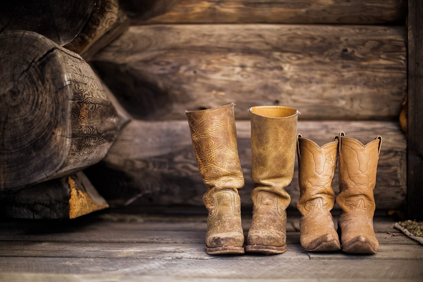 Two pairs of worn cowboy boots on the floor of a log cabin.